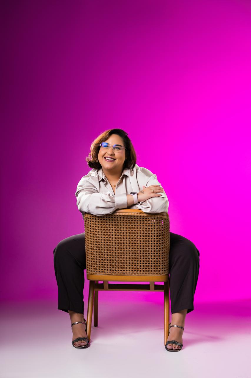 Maria Virginia sentada ao contrário numa cadeira de palhinha, sorrindo, com fundo magenta
