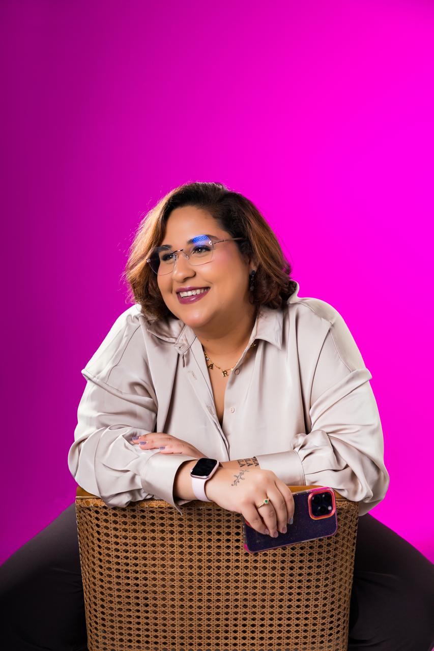Maria Virginia sentada numa cadeira de palhinha, sorrindo, com fundo magenta
